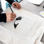 Person painting design on canvas bag with black brush strokes in art studio.